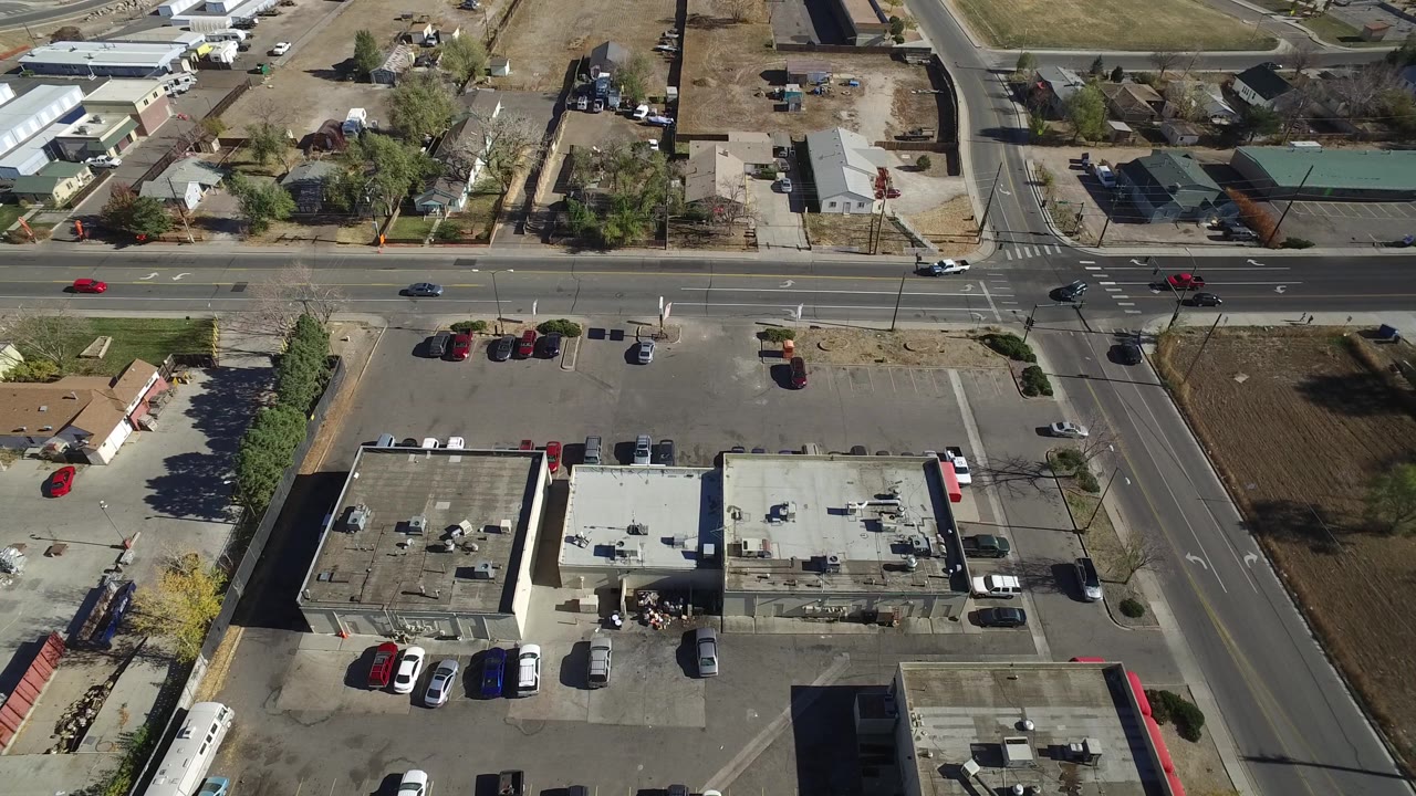 At one second, the Westminster aerial view holds over the intersection with more of the commercial roofs and parking rows visible, plus homes and bare lots beyond, with no visible people.