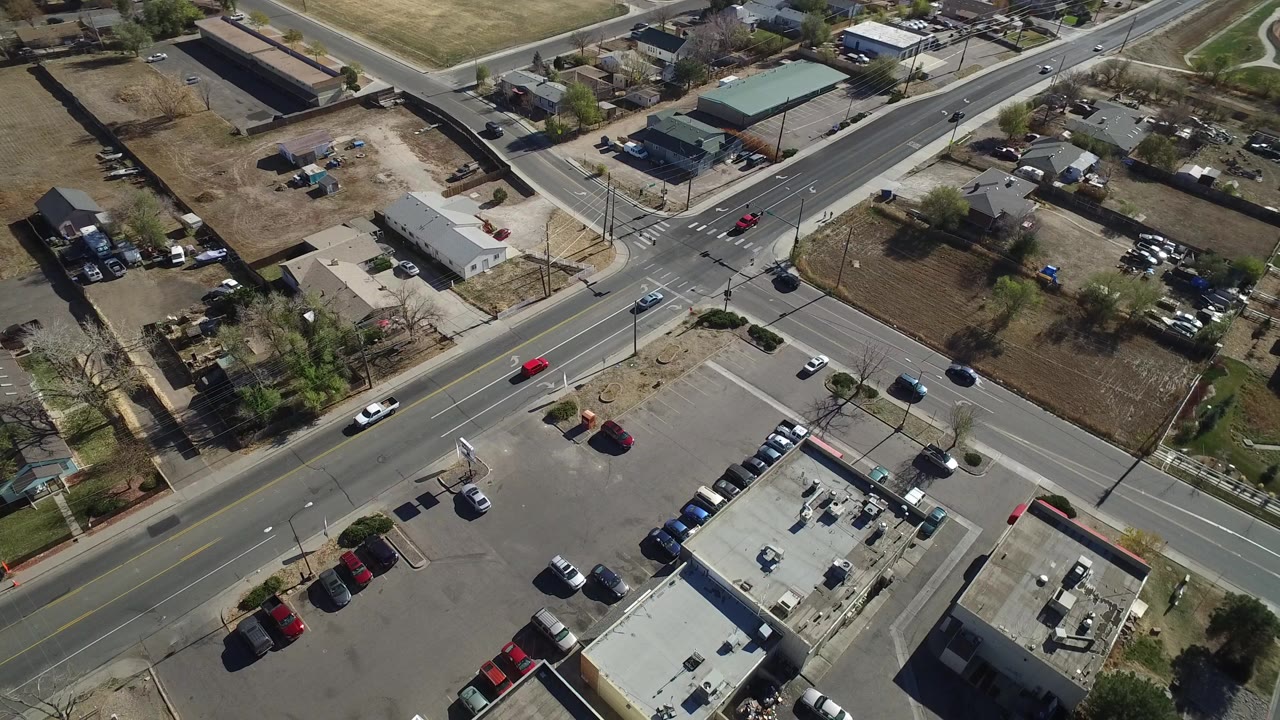 High aerial view over Westminster, Colorado showing a road intersection, low commercial rooftops, parked cars, dry grass areas, and no visible people at the start of the clip.