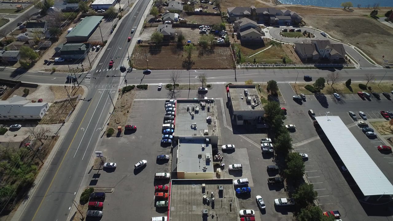 Aerial Westminster, Colorado view with a vertical road beside small businesses, rows of parked cars, a long light-colored roof, residential houses, dry open ground, and tiny people visible near the lower building.