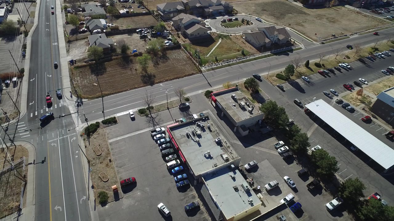 Aerial frame over Westminster, Colorado with a long low building, a corner parking lot, moving road traffic, nearby homes, and no visible people.