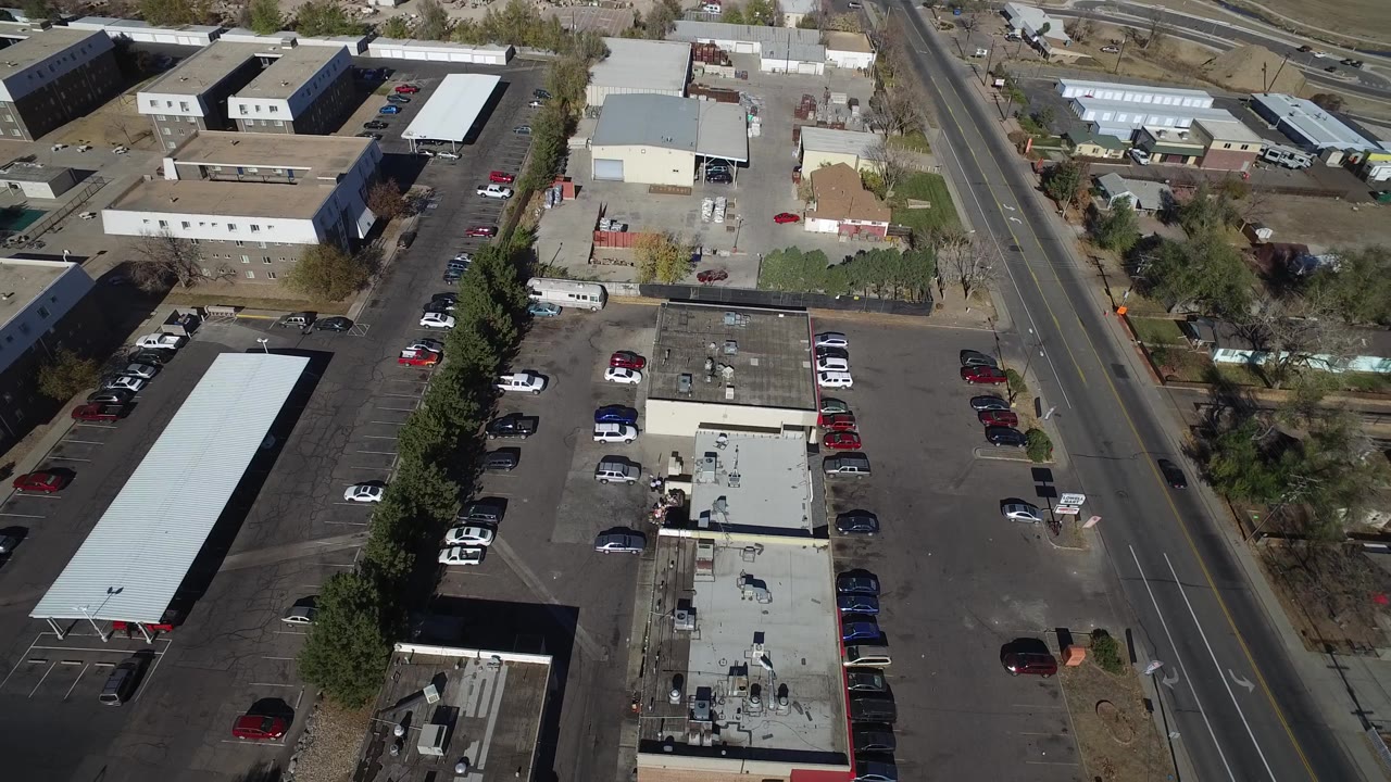 Aerial Westminster, Colorado frame with a long covered structure, flat-roofed buildings, parked cars, a roadside strip, and residential yards in the background, with no visible people.
