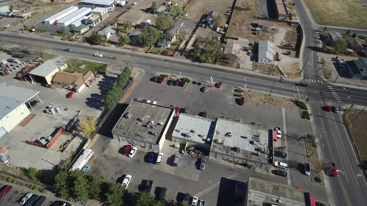Aerial frame in Westminster, Colorado with a main road crossing a side street, flat-roofed businesses, parked vehicles, small residential parcels, and no visible people.