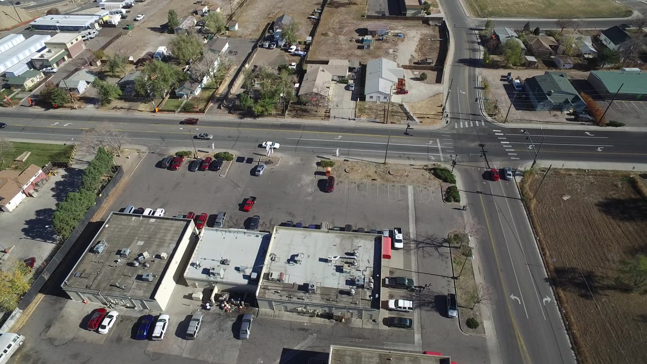 Aerial view over Westminster, Colorado showing a four-way intersection, parked cars, flat-roof commercial buildings, nearby homes, trees, and no visible people.