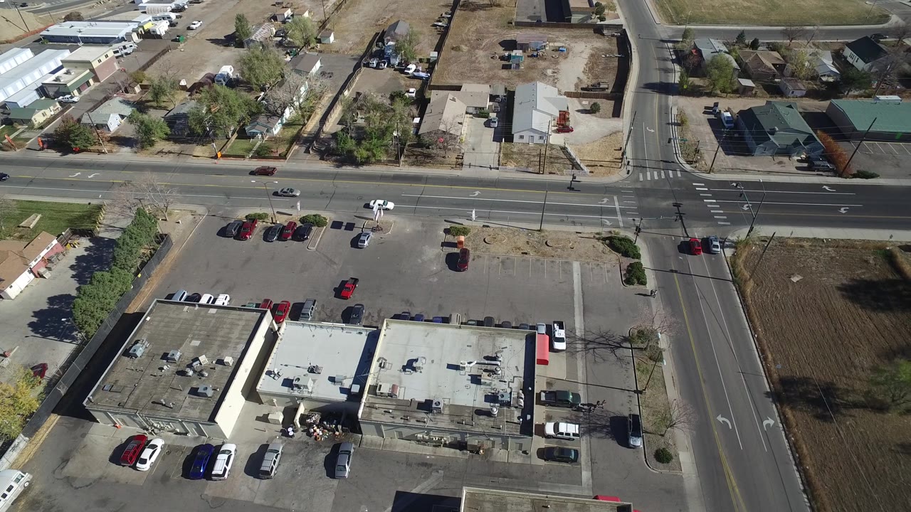Aerial frame in Westminster, Colorado with a white vehicle on the main road, marked lanes, a corner parking lot, low buildings, houses, and no visible people.