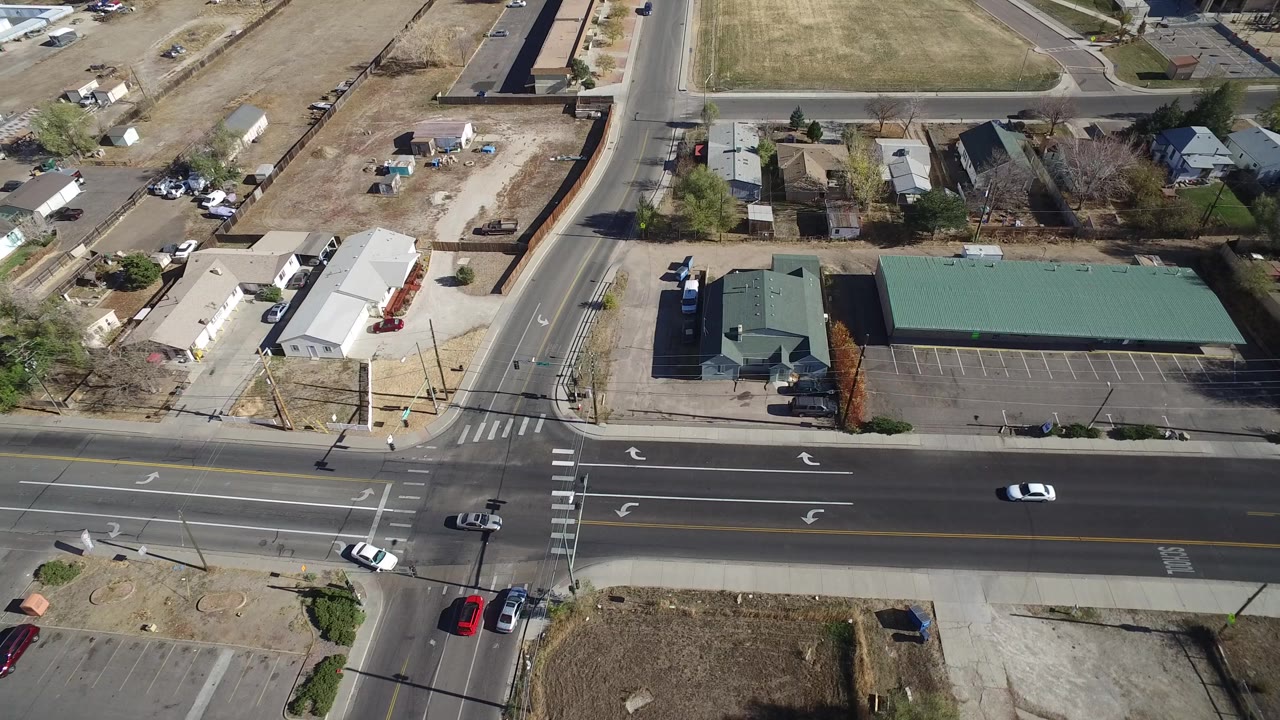 High aerial frame over Westminster, Colorado showing a wide multilane road, a side street, green-roofed commercial buildings, dry lots, homes, light traffic, and no visible people.