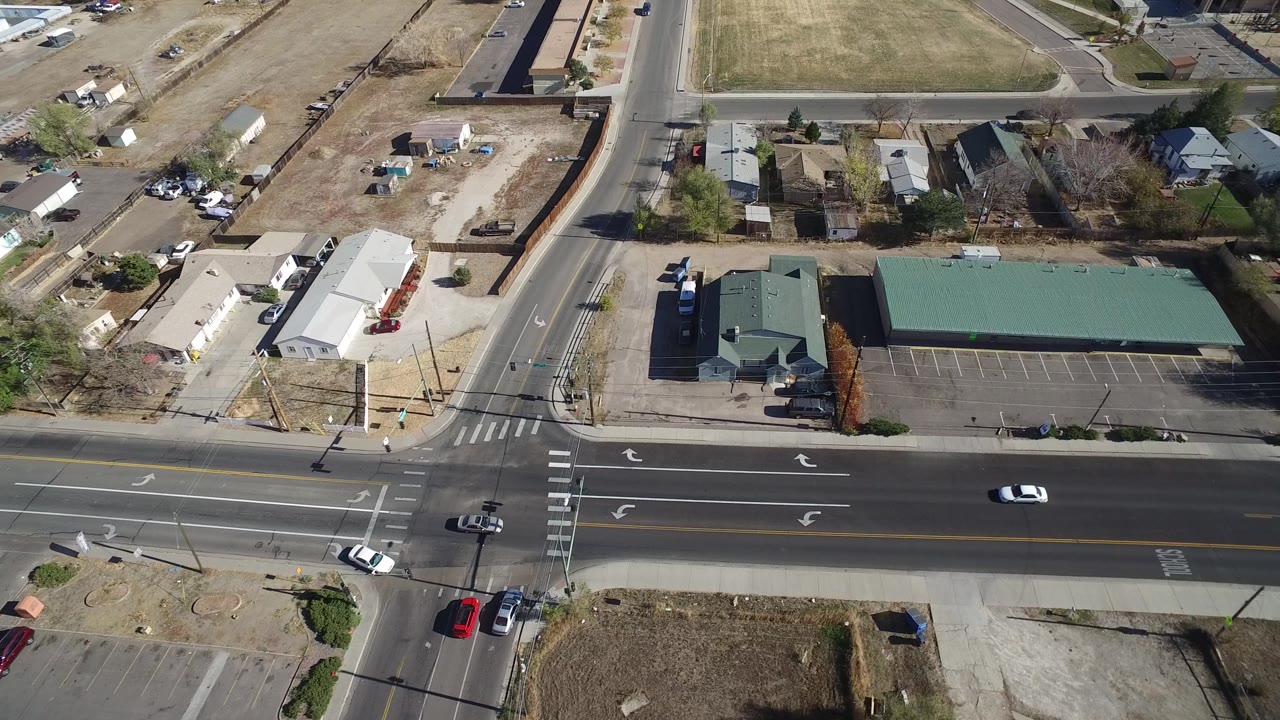 A later aerial frame in Westminster, Colorado keeps the wide road across the foreground while small cars pass near a green-roofed building, parking stalls, dry lots, and no visible people.