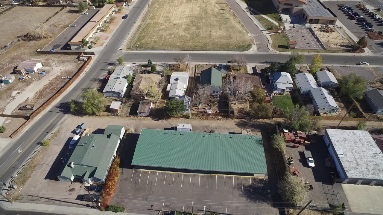 Opening aerial frame over Westminster, Colorado shows a long green-roofed building at the bottom, a mostly empty parking lot, nearby houses, roads, dry grass, and no visible people.