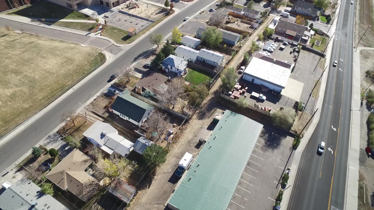 Aerial frame over Westminster, Colorado, with cars on the right-side road, a green-roofed building below, small homes at center, and no clearly visible people.
