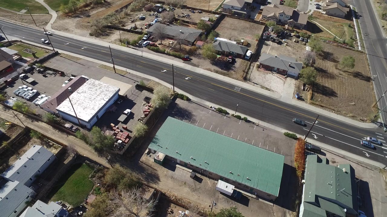Aerial frame over Westminster, Colorado showing a broad road beside green-roofed buildings, a white-roofed structure, parking areas, bare trees, and no visible people.
