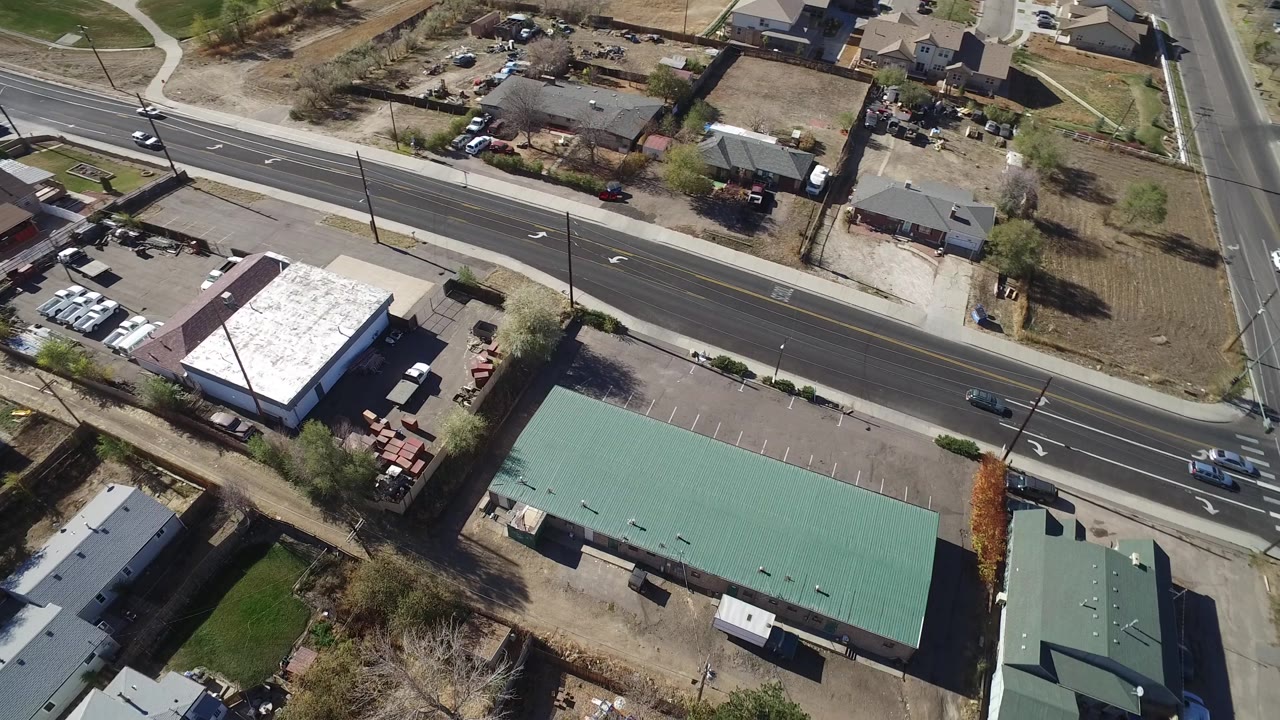 Aerial frame over Westminster, Colorado with the main road running across the image, parking spaces beside green roofs, residential lots beyond, and no visible people.
