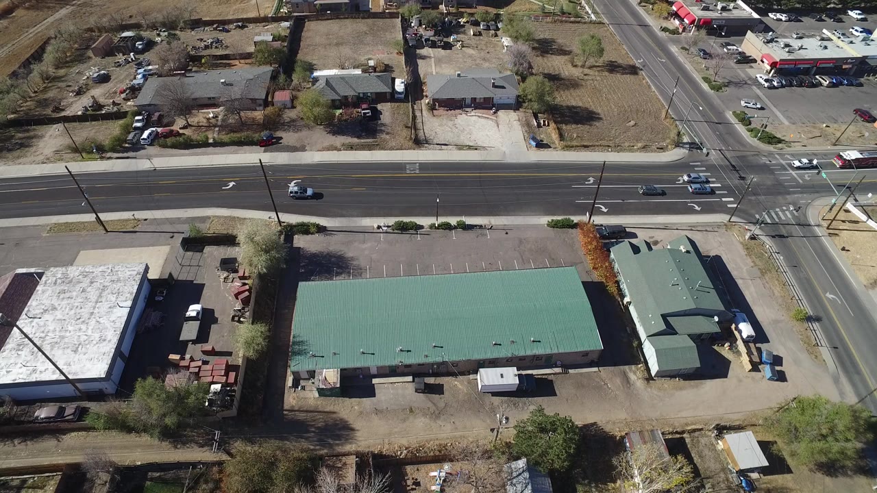 High overhead frame of Westminster, Colorado showing a wide intersection, a long green-roofed commercial building, parking lots, houses, open land, and no visible people.