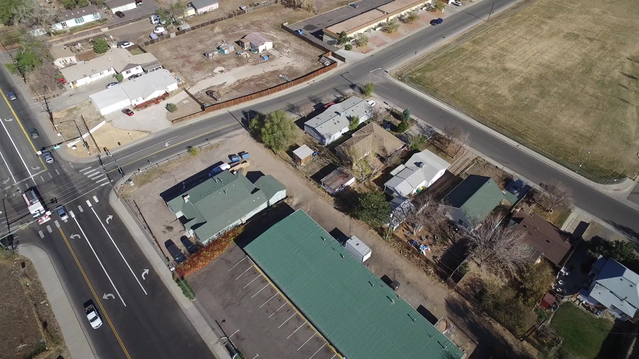 High aerial view over Westminster, Colorado showing a road intersection, green-roofed buildings, parking spaces, private homes, bare trees, dry grass, and no visible people.