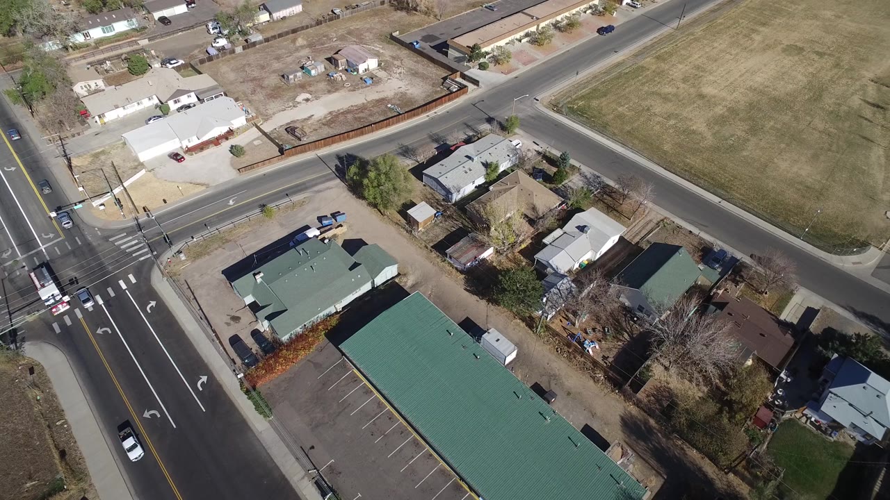 Aerial frame in Westminster, Colorado with a large green-roofed building in the foreground, a diagonal street, nearby houses, parked cars, dry lawns, and no visible people.