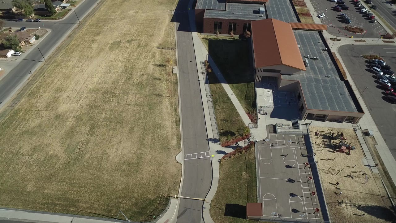 Aerial view of a tan field beside a road, brick building, court, playground, and parking lot.