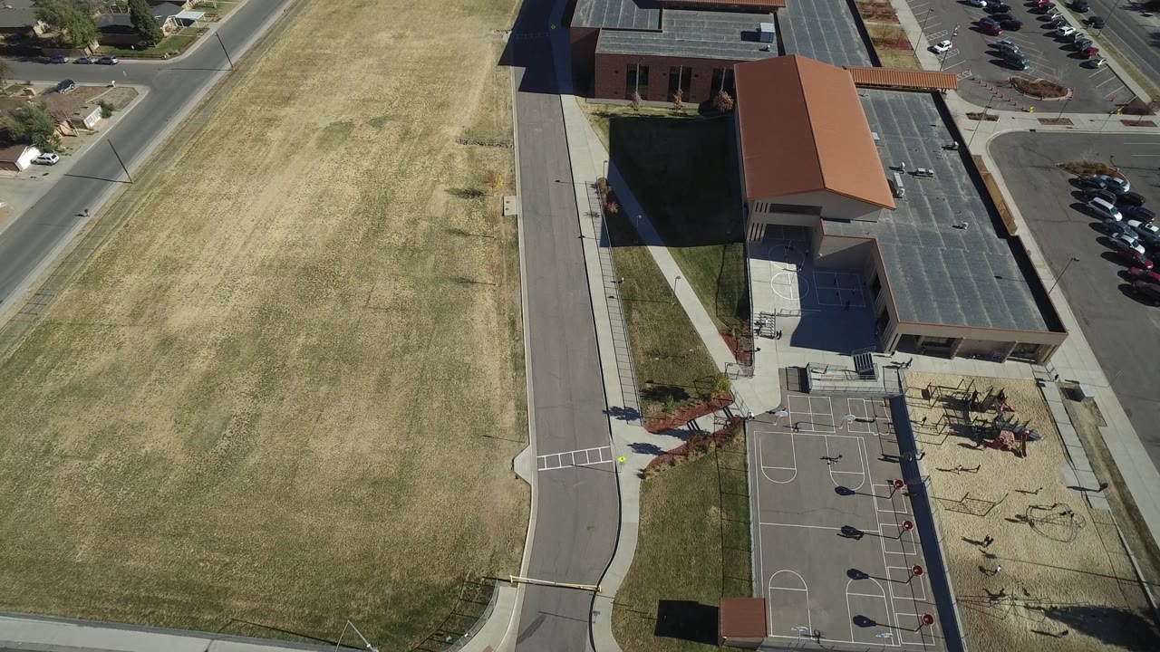 High aerial frame in Westminster, Colorado with the tan field taking the left side, a paved road through the center, and the brick building, court, playground equipment, and parking lot on the right.