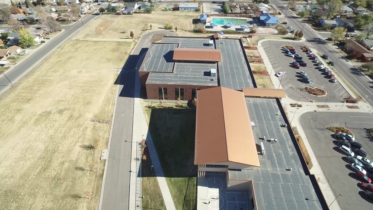 High aerial frame over Westminster, Colorado showing a large tan-roofed building beside a parking lot, a dry grass field to the left, neighborhood streets beyond, and no visible people.