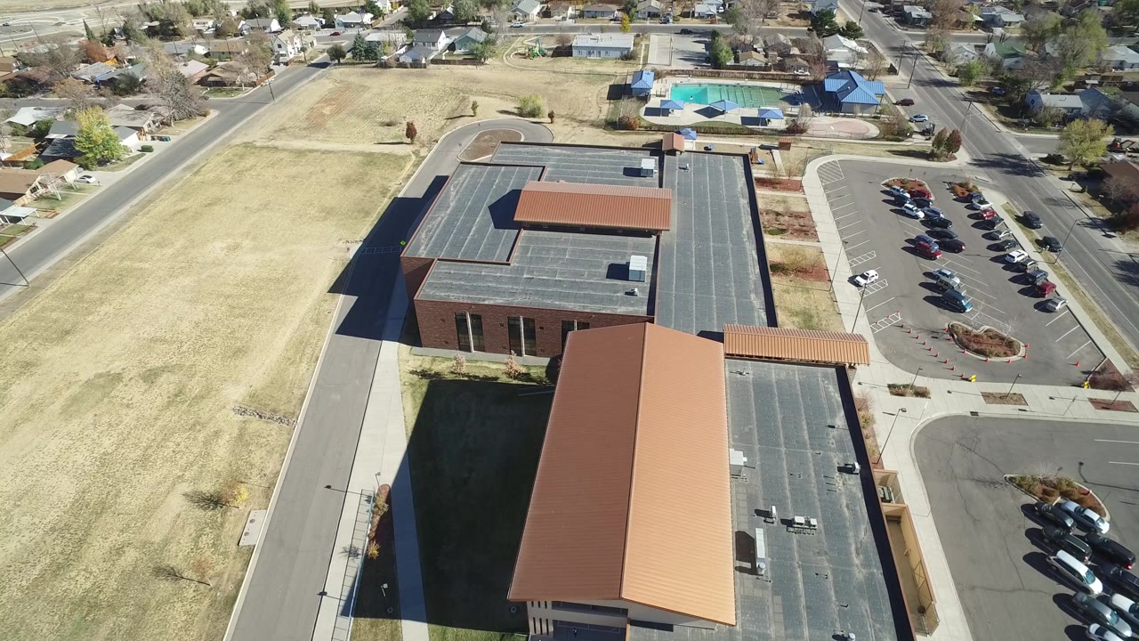 High aerial frame in Westminster, Colorado with the long rust-colored roof centered lower in view, dark-roofed buildings behind it, parking lots to the right, houses beyond the roads, and no visible people.