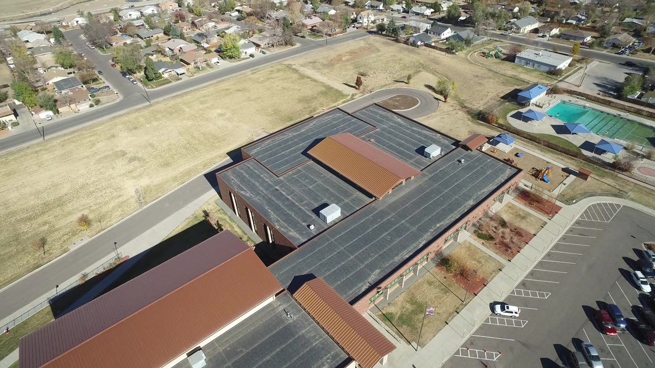 Aerial view of a large building, parking lot, dry field, nearby homes, and pool area.
