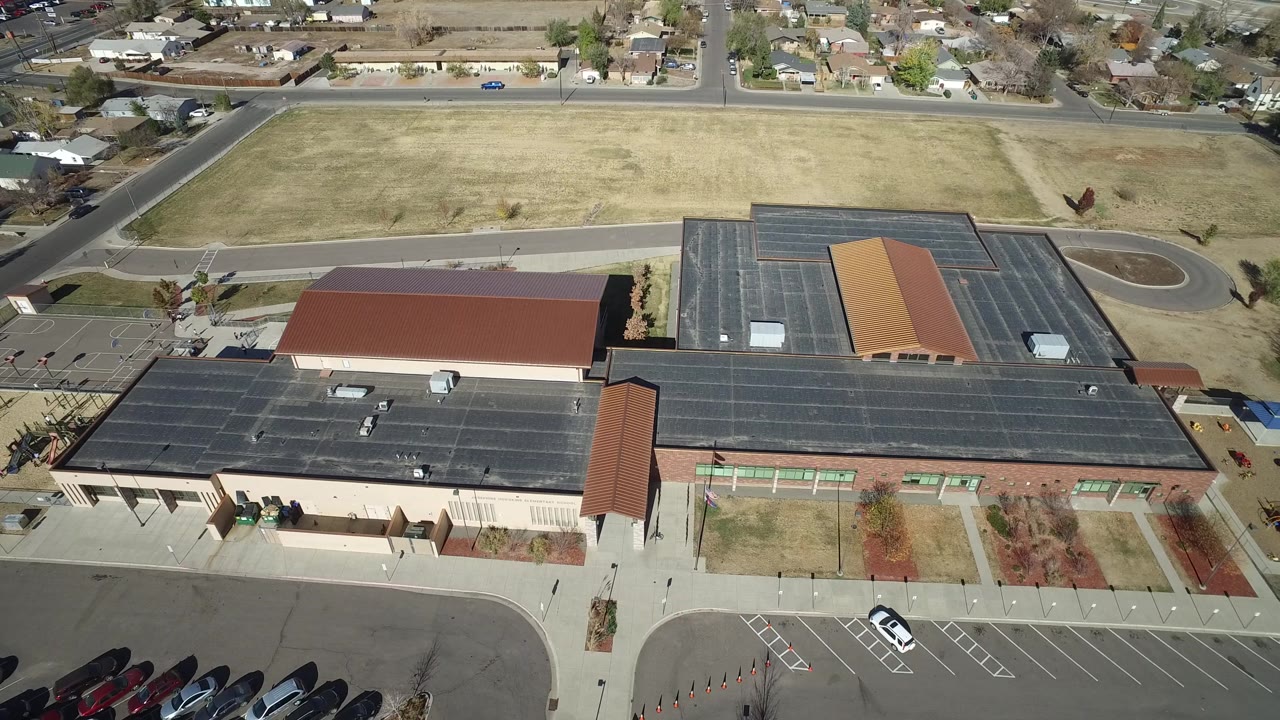 Mid-early aerial frame over the Westminster, Colorado property, with the dark roof stretching across the lower view, parking lanes at front, open grass above, houses at the horizon, and no visible people.