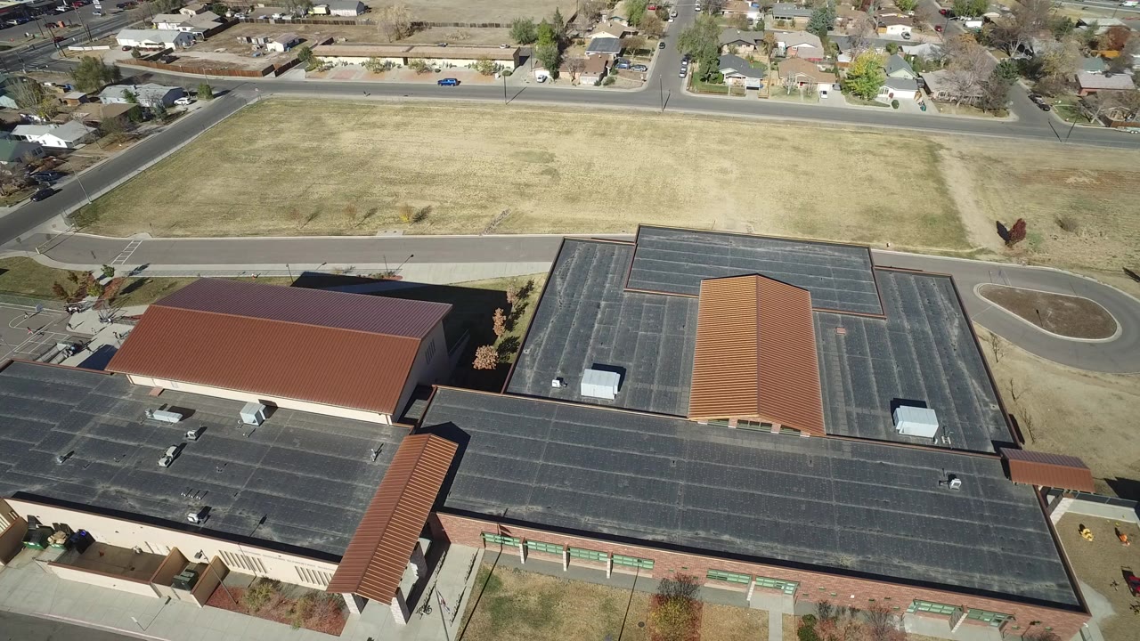 Aerial frame over Westminster, Colorado showing dark-roofed community buildings beside a tan open field, streets, and residential houses, with no visible people.