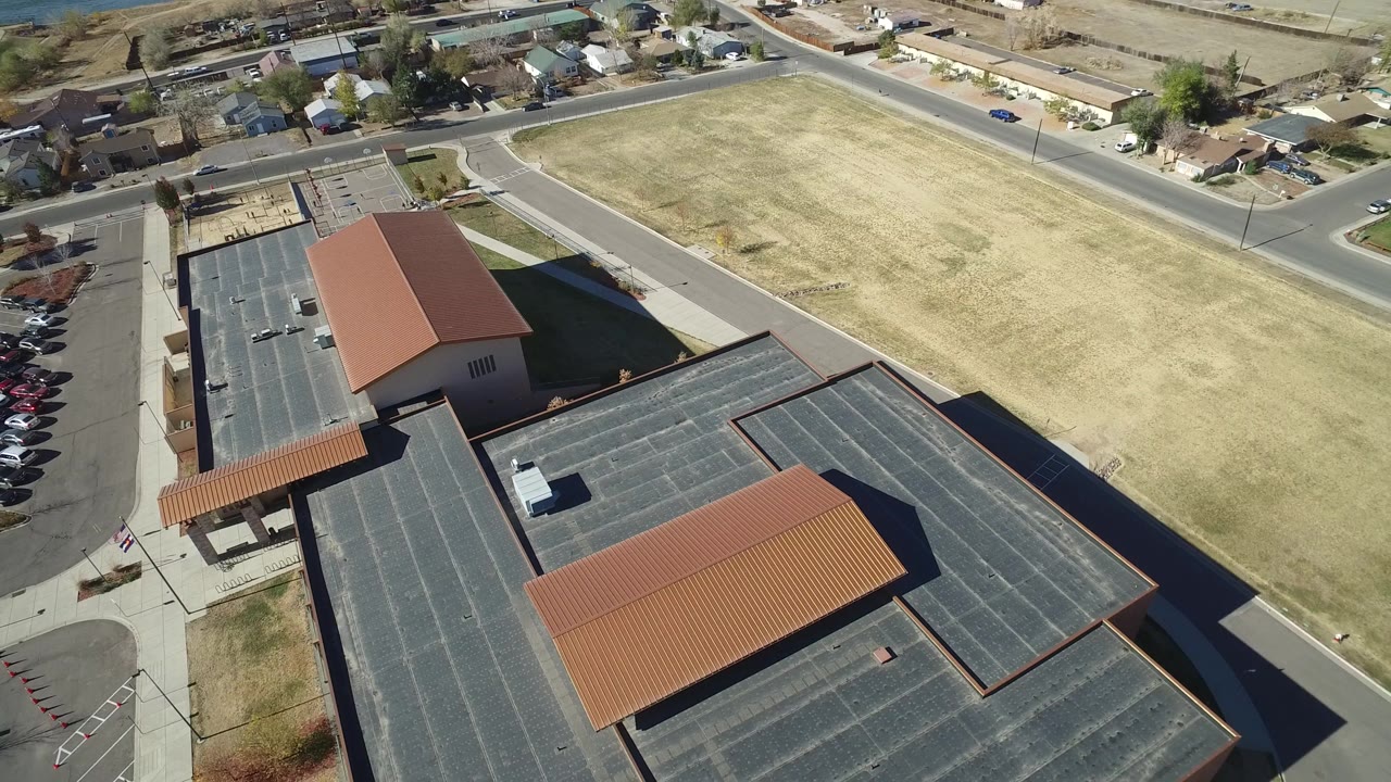 Aerial view one second in shows the Westminster, Colorado building centered lower in frame, with cars along the left parking rows, dry grass to the right, streets above, and no visible people.