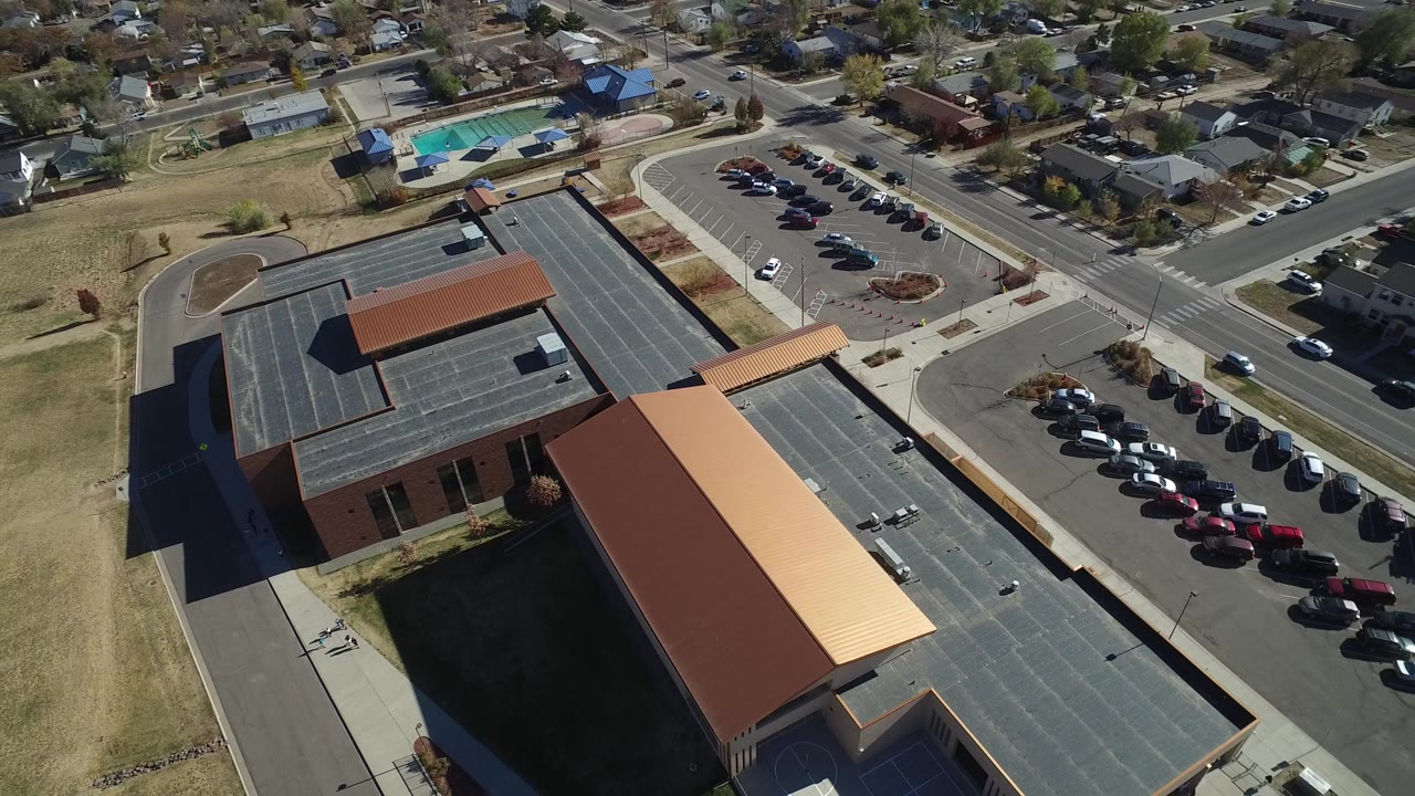 High aerial frame over Westminster, Colorado showing a large red-brown roofed building, striped pavement, a parked-car lot, nearby streets, homes, dry grass, and no visible people.