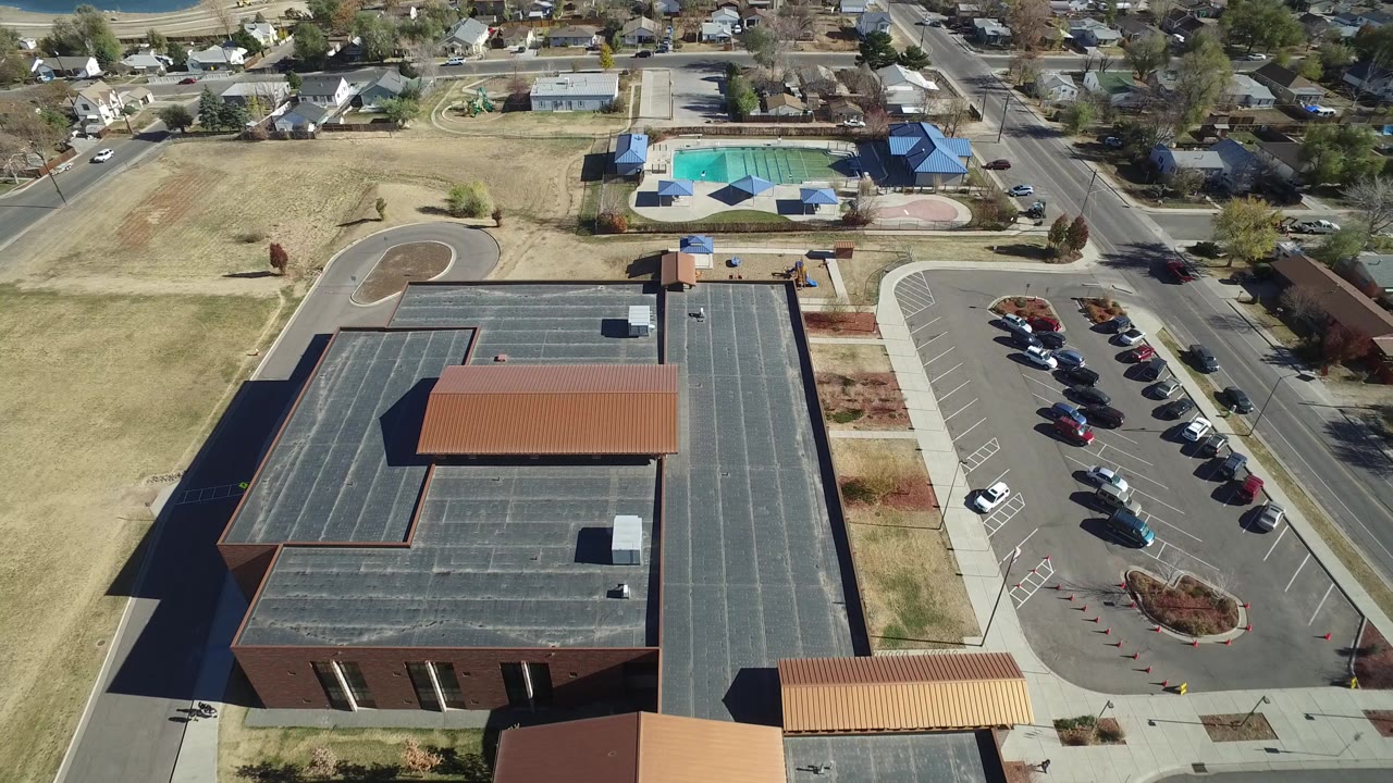 High aerial view in Westminster, Colorado showing a large flat-roofed brick building, parked cars, surrounding homes, dry grass, and a fenced pool area in the distance, with no visible people.