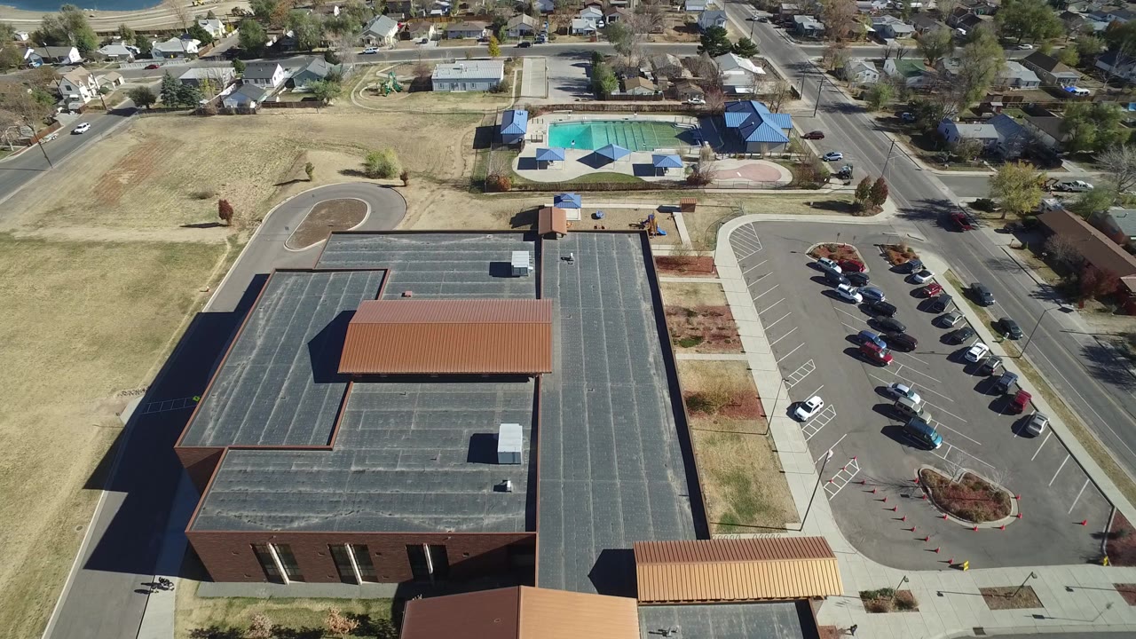 Aerial frame in Westminster, Colorado with the large roof filling the foreground, a parking lot to the right, residential houses beyond, and the blue outdoor pool visible near the top, with no visible people.