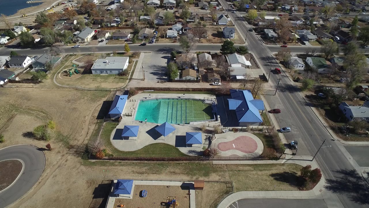 The Westminster, Colorado aerial angle centers the pool deck and blue-roofed structures, with a road on the right, homes beyond it, cars, trees, and tiny visible people near the water.