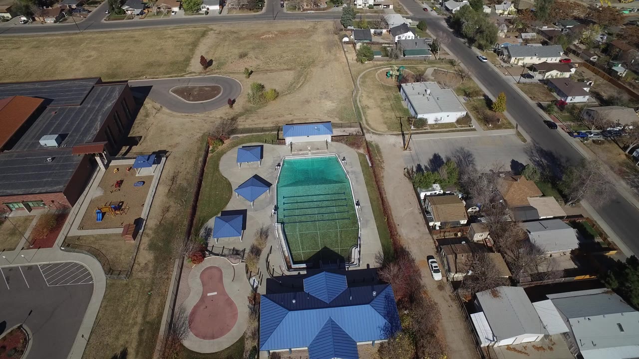 Aerial view of a pool complex with a blue roof, playground, road, and nearby homes.