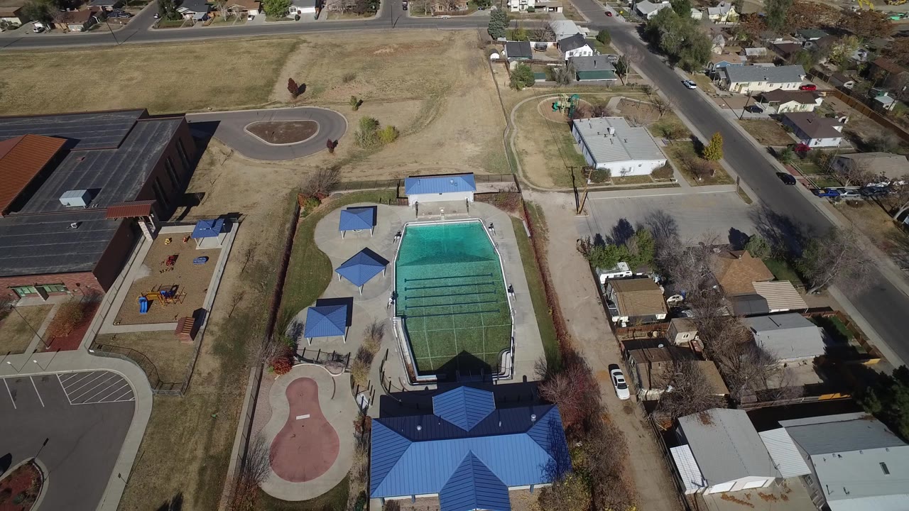 A Westminster, Colorado overhead frame centers the pool complex below a large dark-roofed building, with playground equipment, an open field, a street, and houses visible, with no visible people.