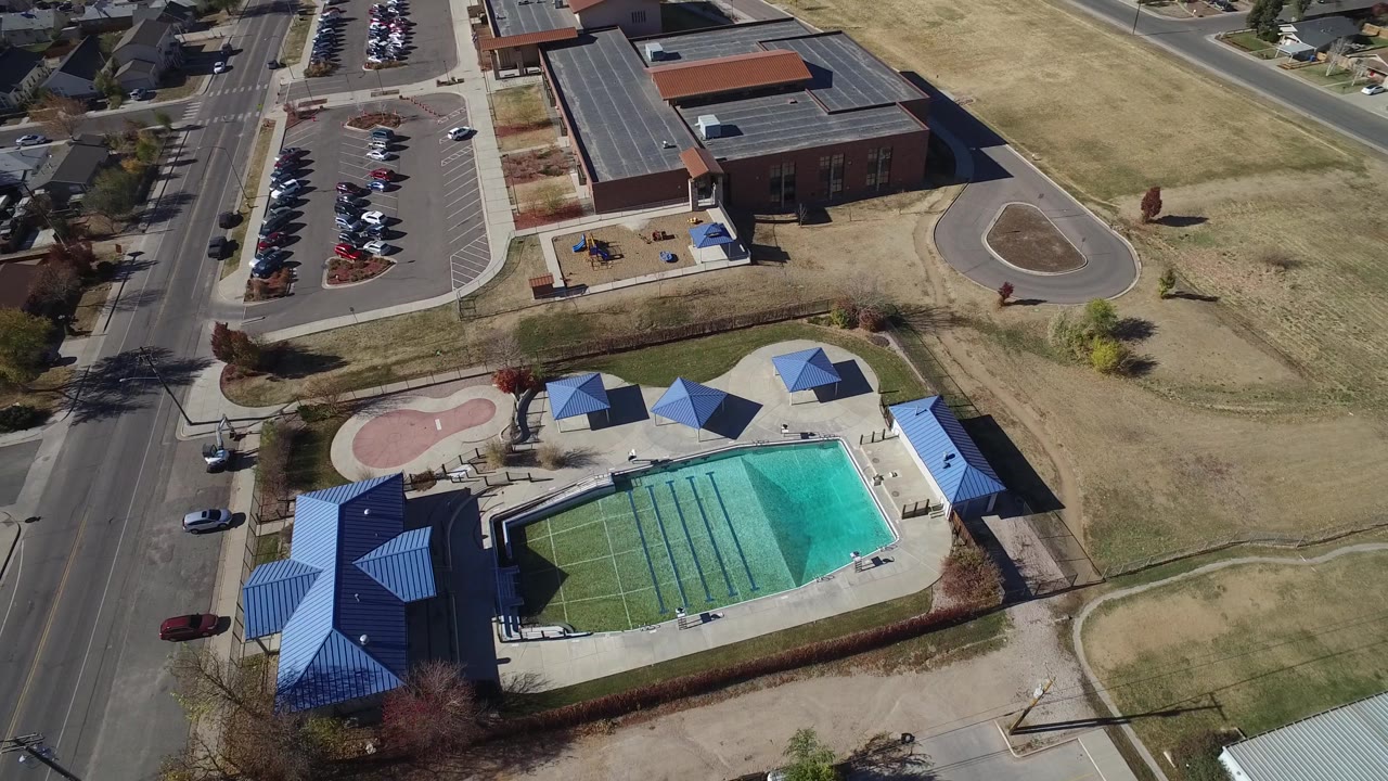 Aerial view in Westminster, Colorado looking down at the same blue-roofed pool facility, with the road and parked cars along the left side, open grass to the right, and no visible people.