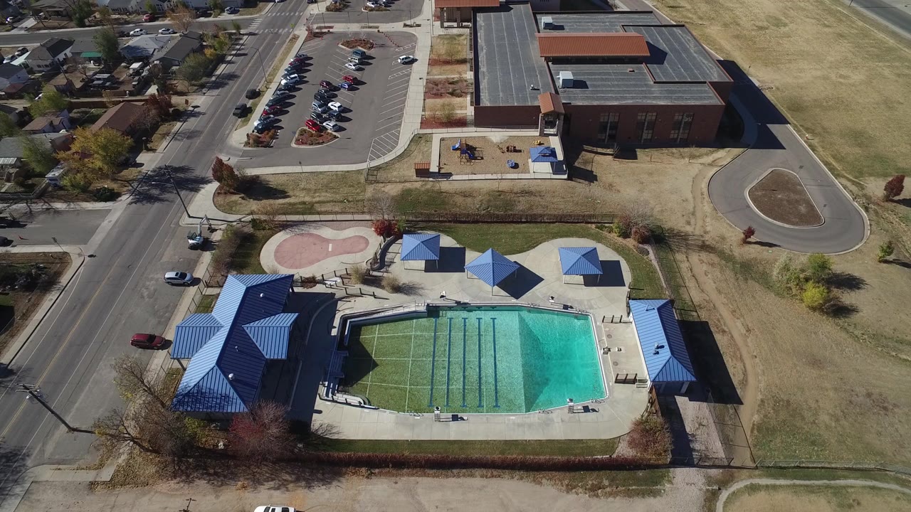 Drone-like overhead view in Westminster, Colorado with the pool centered lower in frame, the road running vertically beside it, parked cars above, and houses in the background, with no visible people.