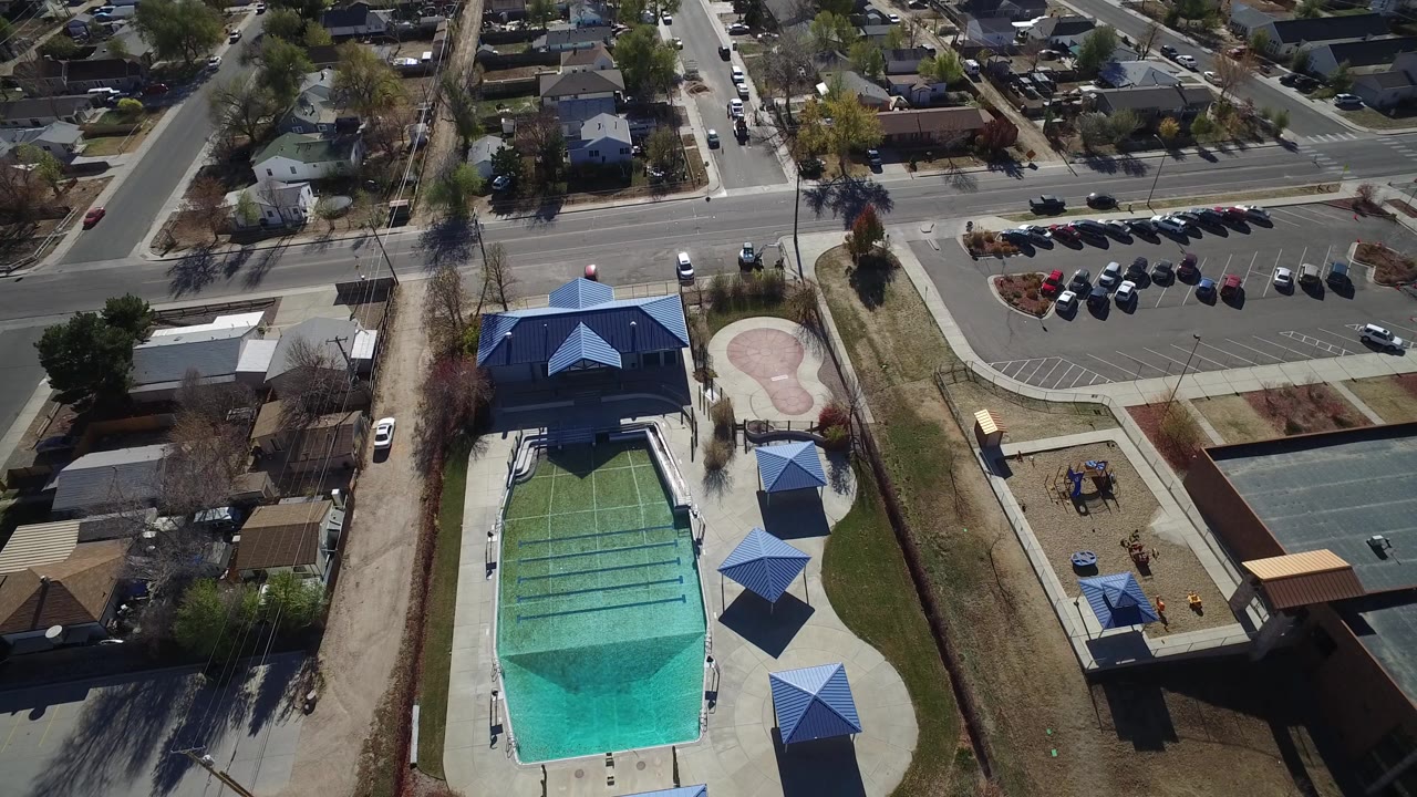 Aerial frame in Westminster, Colorado with the pool more centered below, blue shade roofs along the deck, a curved walkway, a playground to the right, parked cars, and no visible people.
