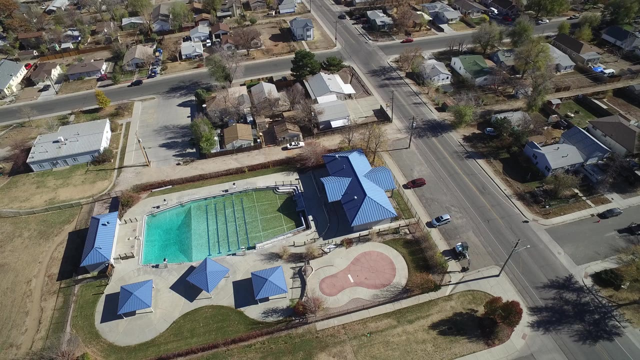 High-angle aerial frame over a Westminster, Colorado outdoor pool complex with blue shade roofs, nearby roads, parked cars, homes, trees, and no visible people.