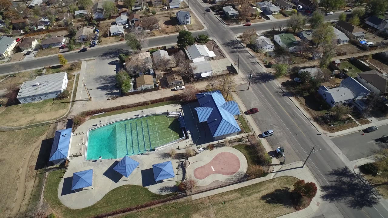 A Westminster, Colorado aerial view centers the turquoise pool and blue-roofed structures, with a broad road at right, houses beyond, dry lawns, and no visible people.