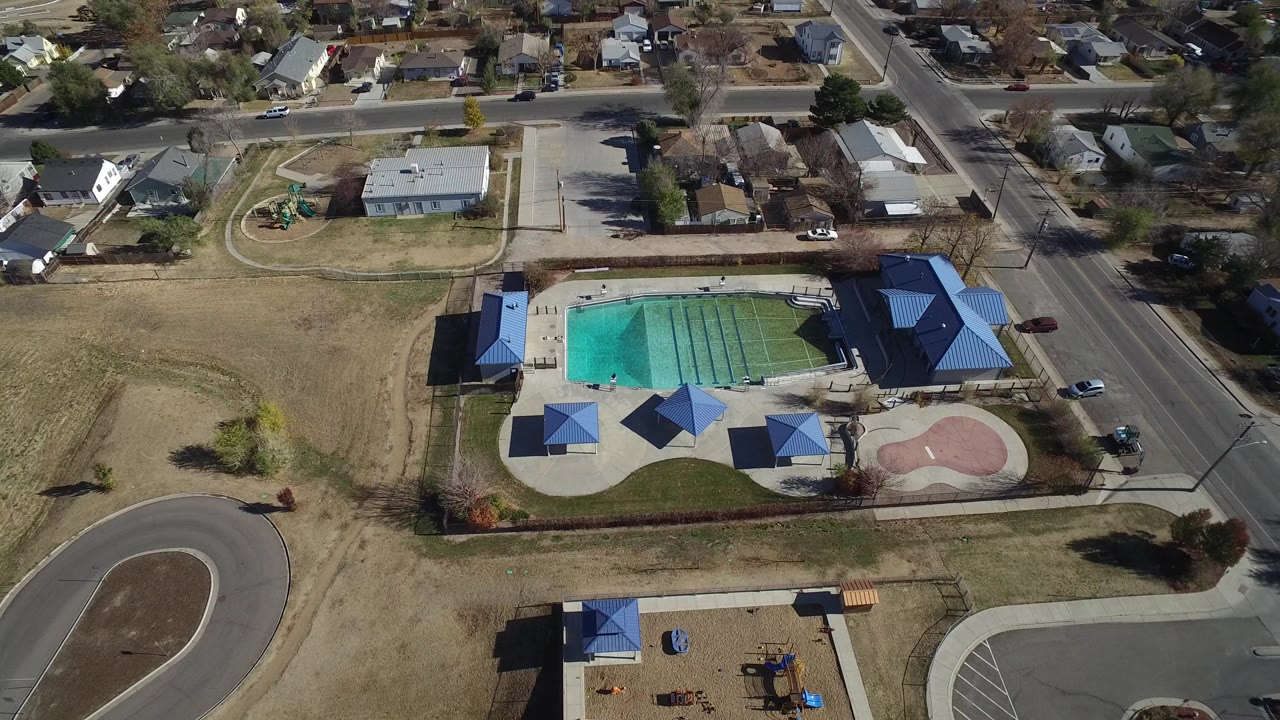 High aerial view over Westminster, Colorado with a turquoise pool, blue shade roofs, curved road, playground, dry grass, parked cars, and nearby houses, with no clearly visible people.