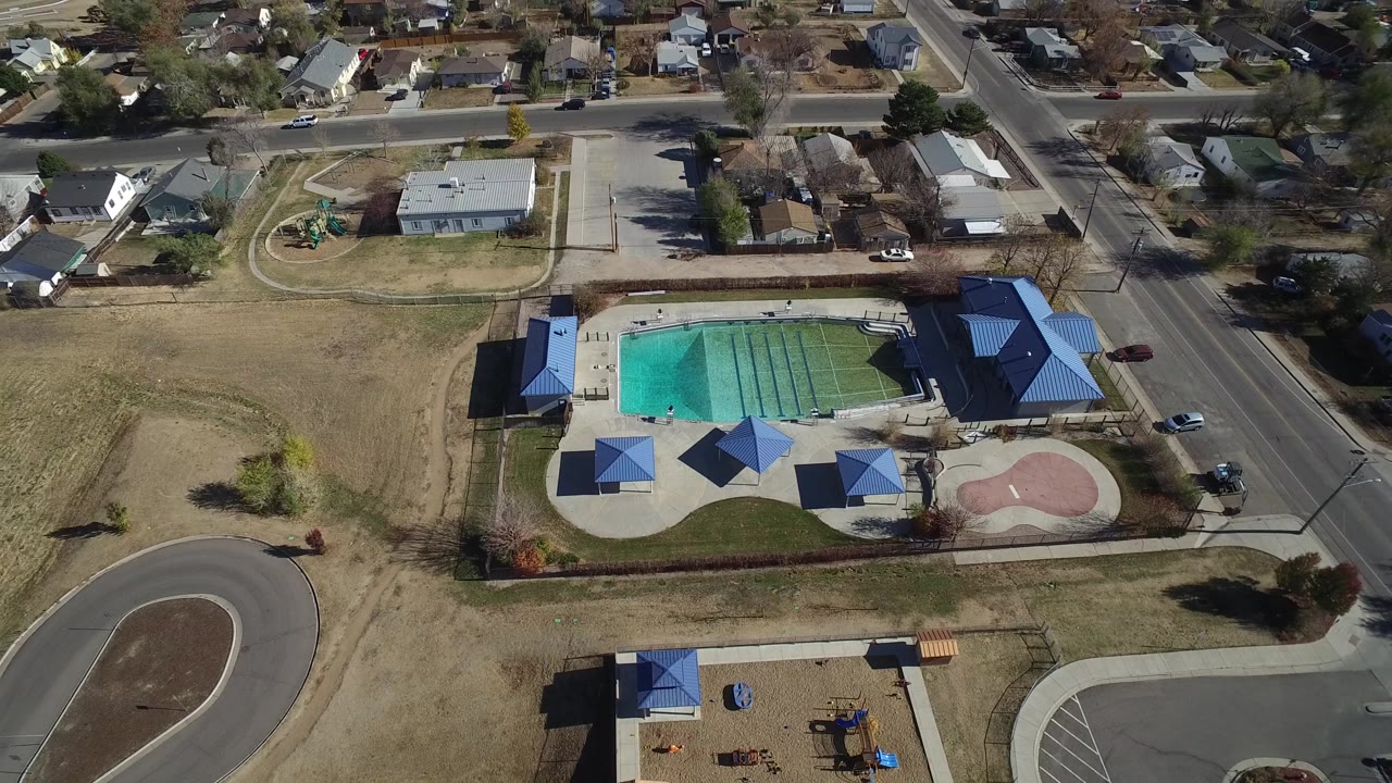 Aerial frame in Westminster, Colorado showing the pool centered near blue-roof shelters, a playground below, a looped road at left, and residential houses beyond, with no clearly visible people.