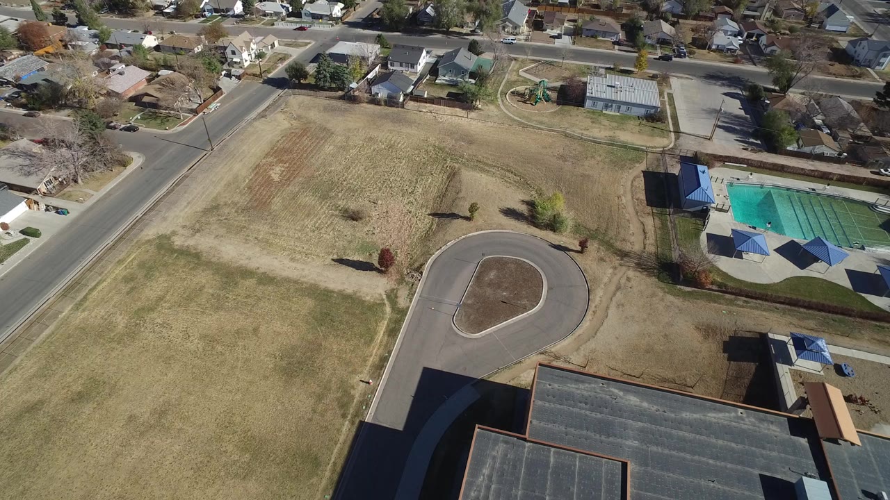High aerial view in Westminster, Colorado showing a tan open field, a curved paved loop, nearby rooftops, neighborhood streets, and no visible people.