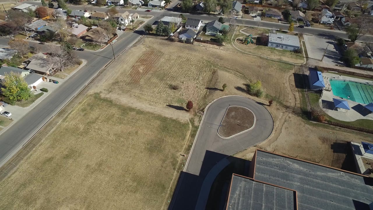 Residential aerial view with dry grass, a loop road, rooftops, and a fenced sports court.