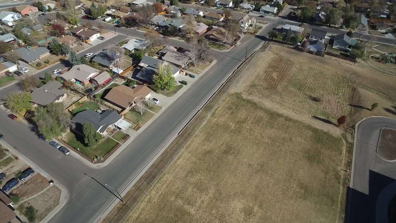 Opening overhead frame in Westminster, Colorado showing a diagonal road intersection, single-family homes, parked cars, a dry open field, and no visible people.