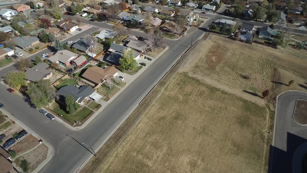 Second overhead frame in Westminster, Colorado with a large dry field on the right, neighborhood homes on the left and top, parked cars, and no visible people.