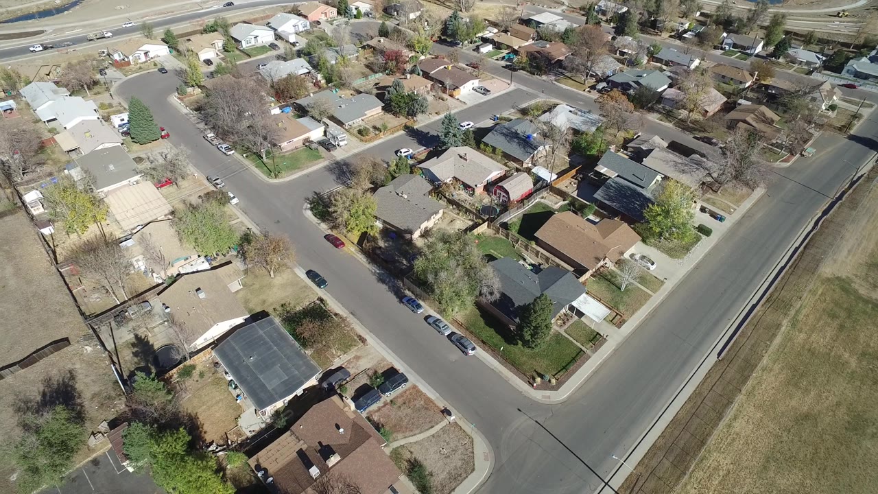 Aerial frame over Westminster, Colorado residential blocks, with rooftops, fenced yards, parked cars along the curb, mixed bare and green trees, and dry open grass at the right edge, with no clearly visible people.