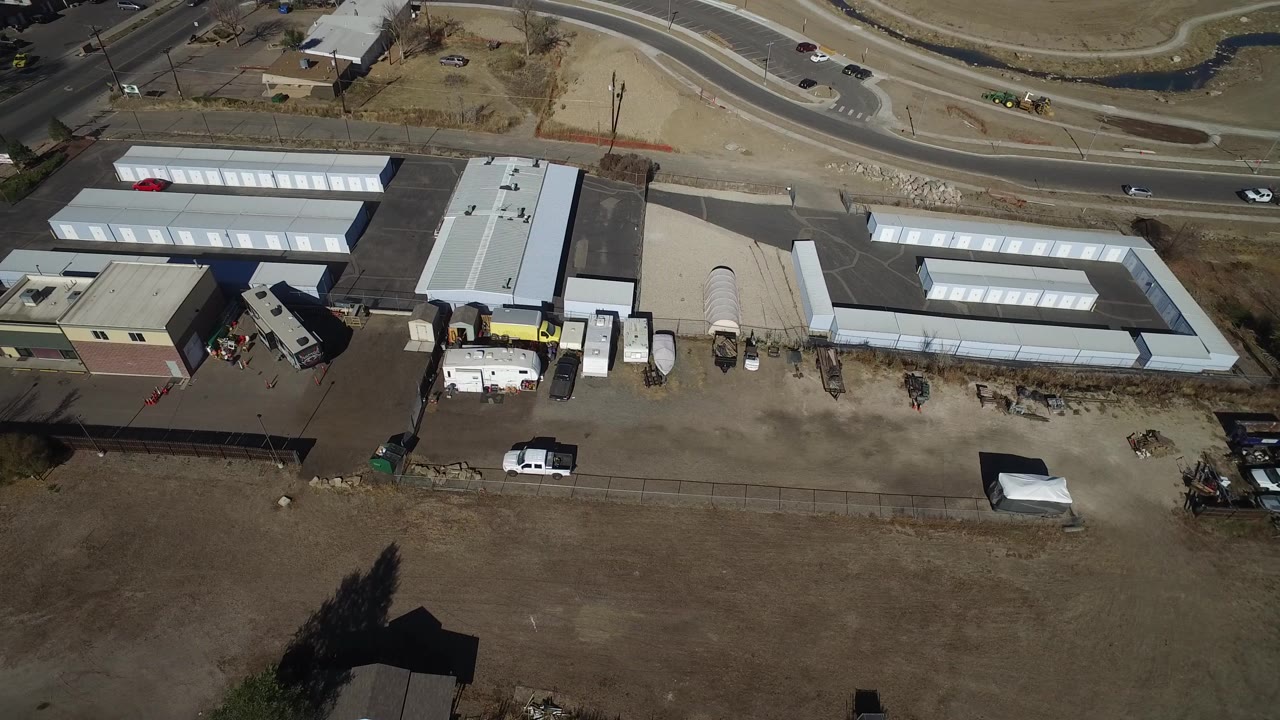 Aerial view in Westminster, Colorado, with storage-unit roofs, asphalt lanes, utility vehicles, trailers, roadside traffic, and no visible people.