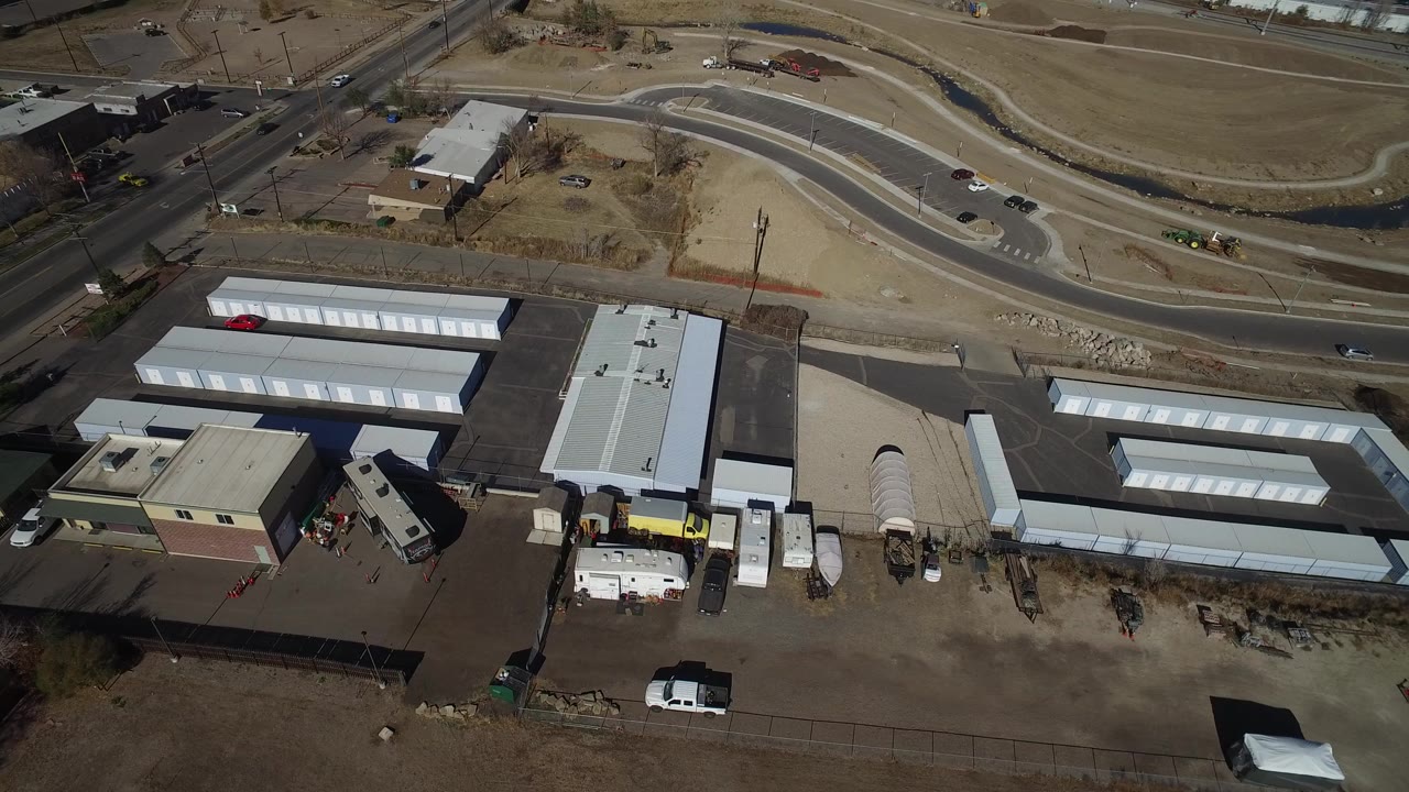 High aerial frame over Westminster, Colorado showing white storage buildings, paved parking areas, a curving road, dry open ground, and no visible people.
