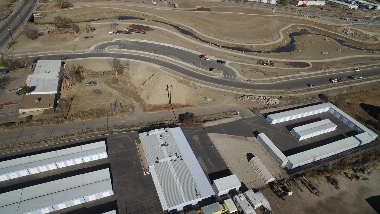 High aerial frame over Westminster, Colorado, with curved roads, bare grass, storage-style rooftops, a parking lot, and no visible people.