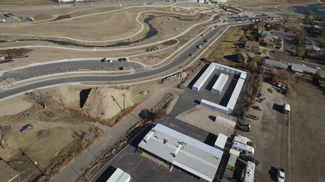 Bird's-eye view in Westminster, Colorado with a road crossing the upper frame, low buildings below, dry lots, scattered cars, a stream bend, and no visible people.