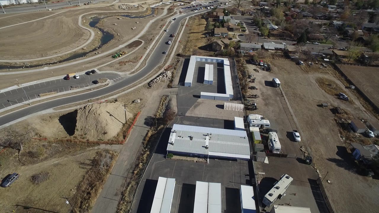 Aerial view in Westminster, Colorado with a warehouse grouping centered lower in frame, storage trailers along the lot, open dirt parcels, a curved road with cars, residential roofs in the background, and no visible people.