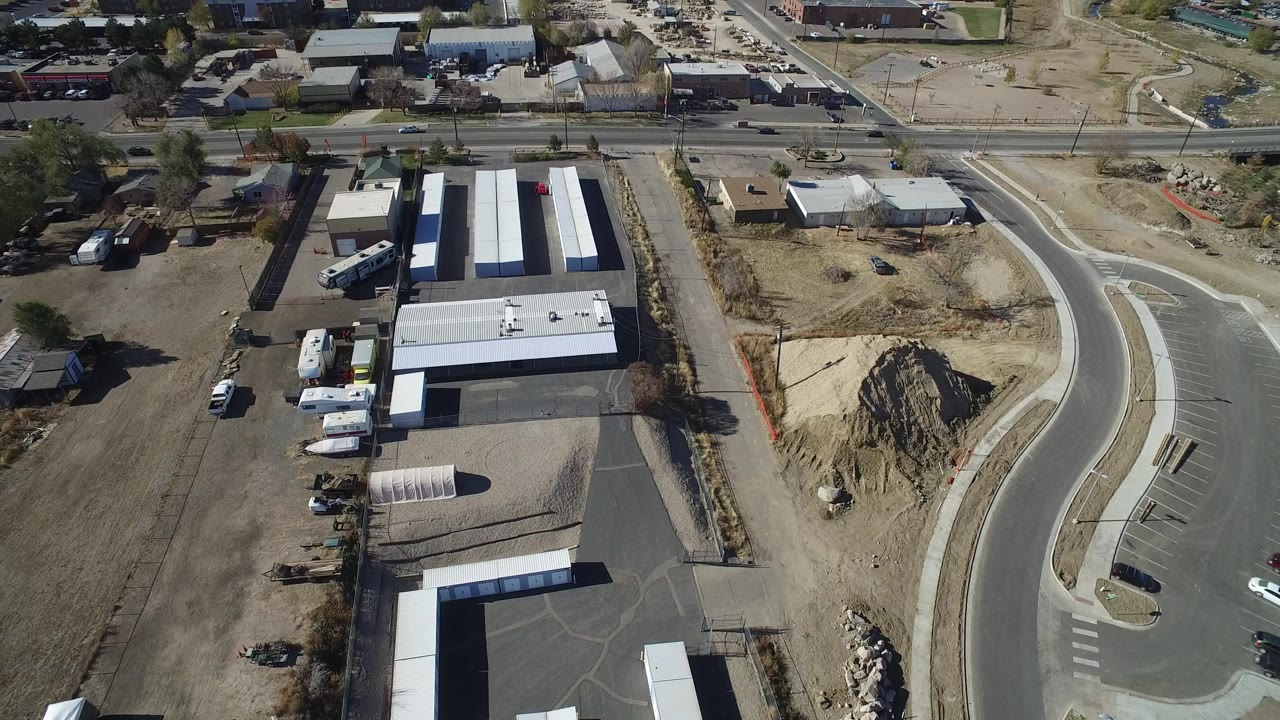 Aerial frame over Westminster, Colorado showing low industrial buildings, parked vehicles, dry dirt lots, a curved road on the right, and no visible people.