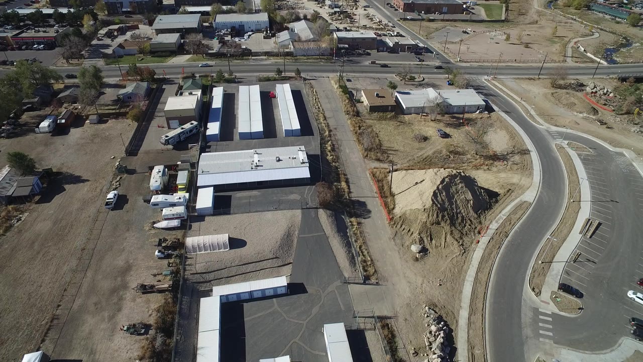 A second aerial view in Westminster, Colorado shows long storage-style buildings, a paved curved roadway, dry lots, parked vehicles, and no visible people.